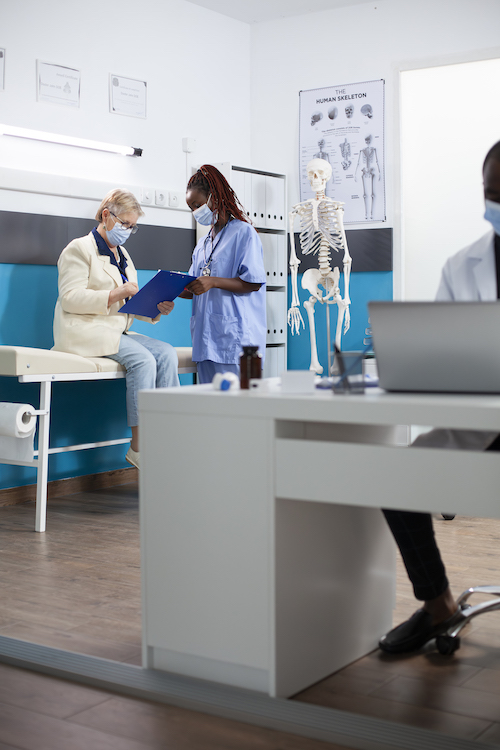 Caucasian senior woman sits in hospital room writing on clipboard as african american nurse offers advice. Female healthcare worker stands attentively guiding elderly patient through medical checkup.